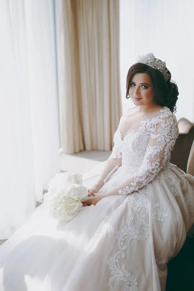 Όμορφη νύφη σε ένα γαμήλιο φόρεμα κάθεται σε μια καρέκλα από το παράθυρο — Φωτογραφία Αρχείου