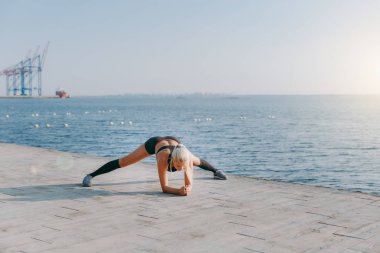 Eğitim ve gündoğumu denize, aşağı eğilerek egzersizleri yapıyor siyah giysili uzun sarı saçlı genç güzel atletik kız