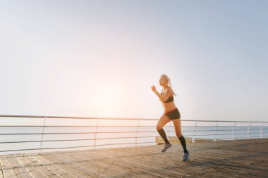 Güneş doğarken denizin üzerinde çalışan siyah giysili uzun sarı saçlı genç güzel atletik kız