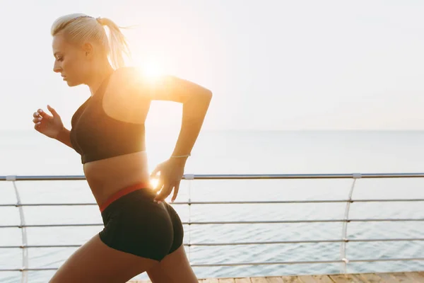 Young beautiful athletic girl with long blond hair in black clothes running at sunrise over the sea