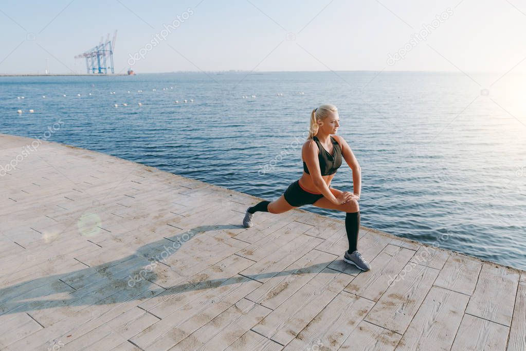 Young beautiful athletic girl with long blond hair in black clothes doing stretching at sunrise over the sea