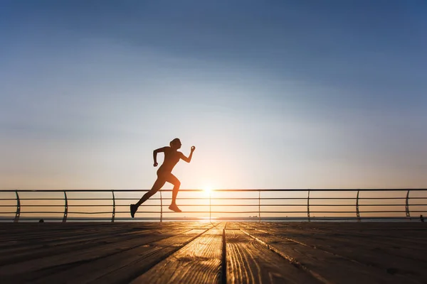 Silueta de una joven hermosa atlética con el pelo largo y rubio en ropa negra corriendo al amanecer sobre el mar — Foto de Stock