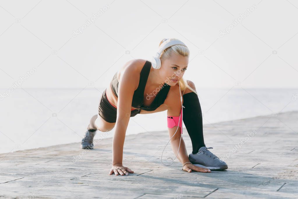 Young beautiful athletic girl with long blond hair in headphones and mobile phone on her hand listening to music and doing stretching at sunrise over the sea