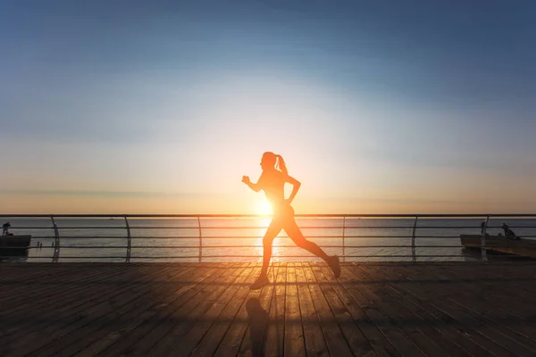 Silueta de una joven hermosa atlética con el pelo largo y rubio en ropa negra corriendo al amanecer sobre el mar — Foto de Stock
