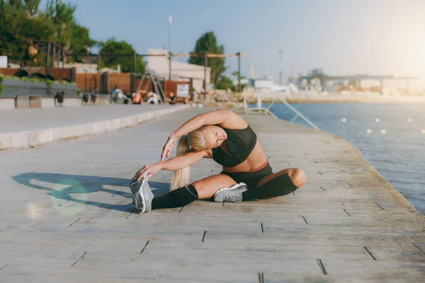 Joven chica atlética hermosa con el pelo largo y rubio en el entrenamiento de ropa negra y haciendo ejercicios al amanecer sobre el mar — Foto de Stock