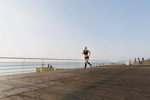 Young beautiful athletic girl with long blond hair in black clothes running at sunrise over the sea