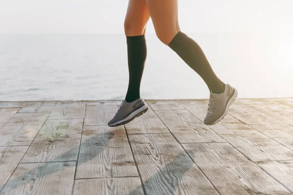 Patas de niña en polainas negras y zapatillas grises, que corre por la mañana al amanecer cerca del mar — Foto de Stock