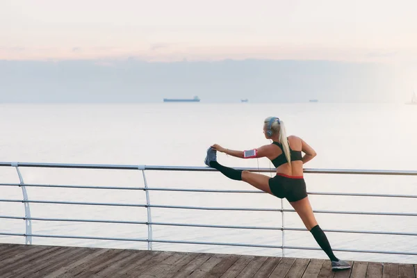 Joven chica atlética hermosa con el pelo largo y rubio en los auriculares escuchando música y haciendo estiramiento al amanecer sobre el mar, vista trasera — Foto de Stock