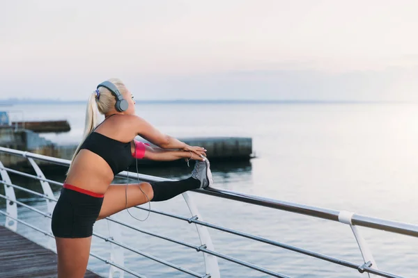 Joven chica atlética hermosa con el pelo largo y rubio en los auriculares escuchando música y haciendo estiramiento al amanecer sobre el mar — Foto de Stock