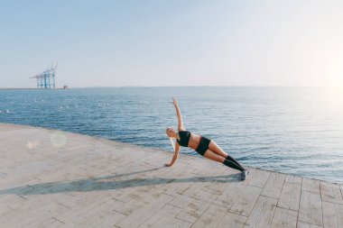 Eğitim ve egzersizleri, yapıyor siyah giysili uzun sarı saçlı genç güzel atletik kız kolunu kadar güneş doğarken denizin üzerinde yükselterek