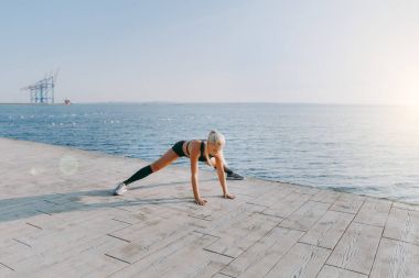 Eğitim ve gündoğumu denize, aşağı eğilerek egzersizleri yapıyor siyah giysili uzun sarı saçlı genç güzel atletik kız