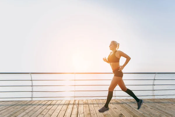 Joven chica atlética hermosa con el pelo largo y rubio en ropa negra corriendo al amanecer sobre el mar — Foto de Stock