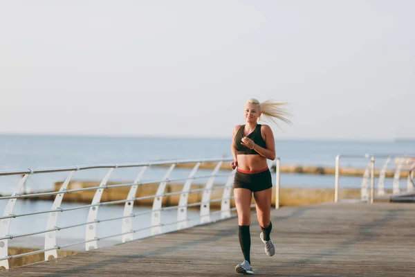 Joven chica atlética hermosa con el pelo largo y rubio en ropa negra corriendo al amanecer sobre el mar — Foto de Stock