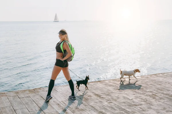 Όμορφη αθλητική κοπέλα με μακριά ξανθά μαλλιά, μαύρα ρούχα, και δύο από τα σκυλιά της κάνει σπορ με την Ανατολή από τη θάλασσα — Φωτογραφία Αρχείου
