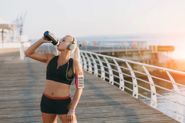 Joven chica atlética hermosa con el pelo largo y rubio en los auriculares bebe agua de una botella al amanecer junto al mar — Foto de Stock