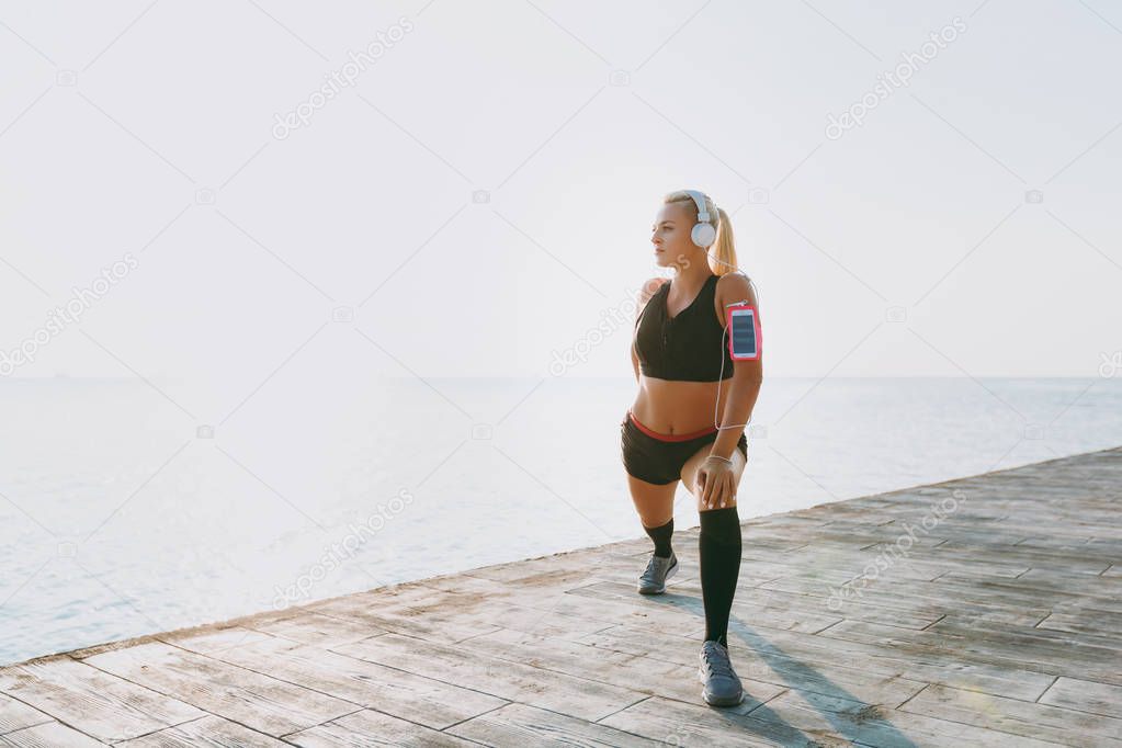 Young beautiful athletic girl with long blond hair in headphones and mobile phone on her hand listening to music and doing stretching at sunrise over the sea
