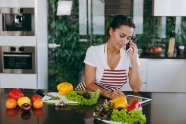 Mutlu kadın cep telefonu üzerinde konuşurken ve tarifi mutfakta tablet bakarak önlük. Sebze salatası. Diyet kavramı. Sağlıklı bir yaşam tarzı. Evde yemek. Yemek hazırlamak.