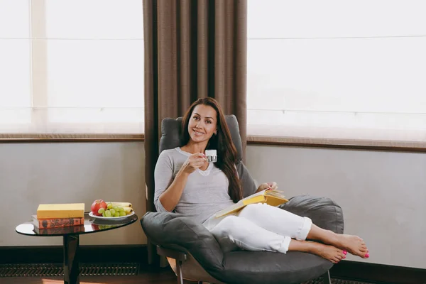 Bella giovane donna felice a casa seduta sulla sedia moderna davanti alla finestra, rilassarsi nel suo soggiorno, leggere libri e bere caffè o tè — Foto Stock