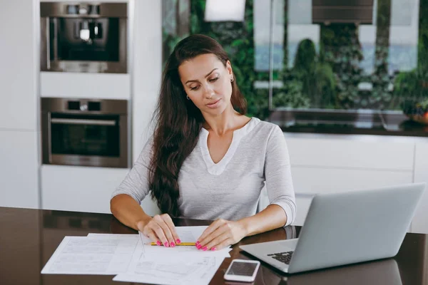 Привлекательная молодая современная деловая женщина, работающая с документами и ноутбуком на кухне дома — стоковое фото