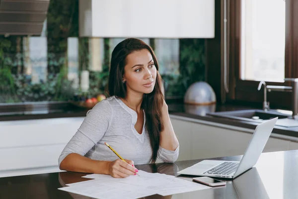 Attractive young pensive business woman working with documents and laptop in the kitchen at home — Stock Photo, Image