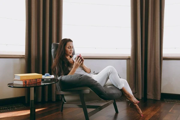 Молодая женщина дома сидит на современном стуле перед окном, отдыхает в своей гостиной и пьет кофе или чай — стоковое фото
