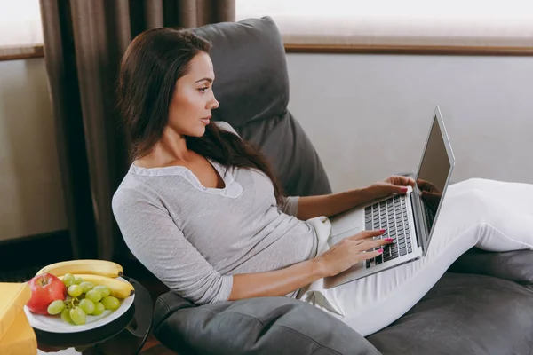 Mulher bonita em casa sentada na cadeira moderna na frente da janela, relaxando em sua sala de estar e trabalhando com laptop — Fotografia de Stock