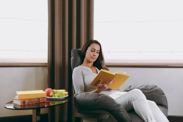 Bella giovane donna a casa seduta sulla sedia moderna davanti alla finestra, rilassante nel suo soggiorno e libro di lettura — Foto Stock