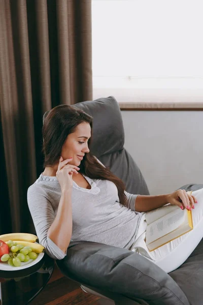 Молодая женщина дома сидит на современном стуле перед окном, отдыхает в гостиной и читает книгу — стоковое фото