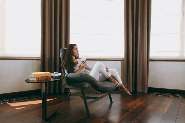 Mulher bonita em casa sentada na cadeira moderna na frente da janela, relaxando em sua sala de estar e bebendo café ou chá — Fotografia de Stock