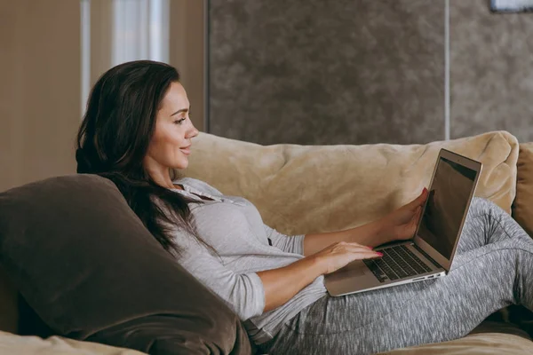 Mulher bonita em casa deitada no sofá, relaxando em sua sala de estar e trabalhando com laptop — Fotografia de Stock