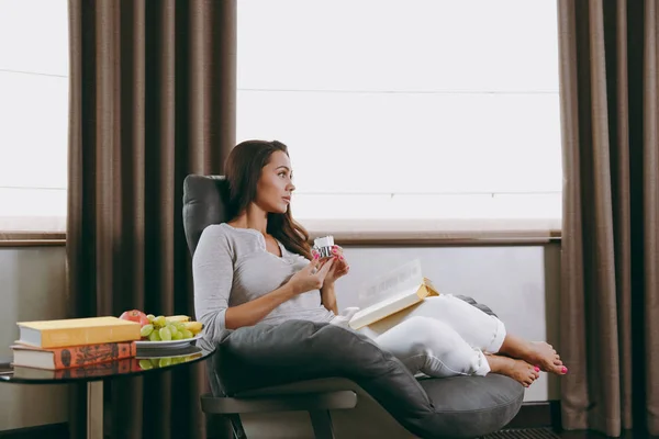 Молодая женщина дома сидит на современном стуле перед окном, отдыхает в своей гостиной, читает книгу и пьет кофе или чай — стоковое фото