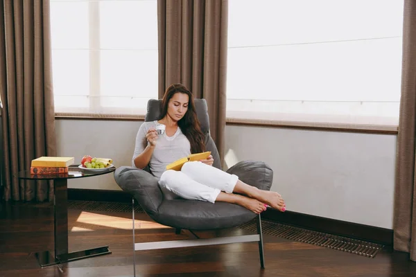 Bella giovane donna a casa seduta sulla sedia moderna davanti alla finestra, rilassarsi nel suo soggiorno, leggere libri e bere caffè o tè — Foto Stock