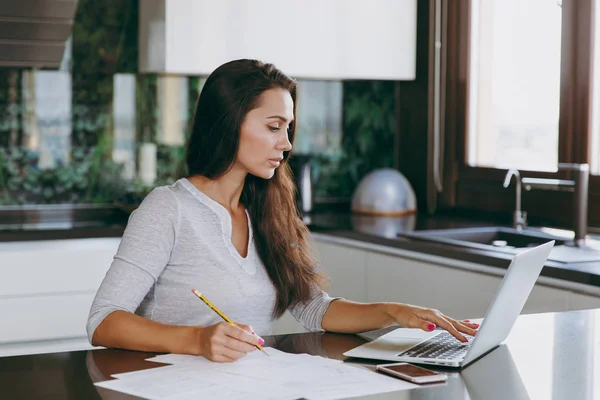 Привлекательная молодая современная деловая женщина, работающая с документами и ноутбуком на кухне дома — стоковое фото