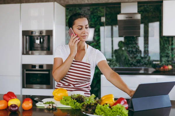 Giovane donna felice in grembiule parlando al telefono cellulare e guardando la ricetta in computer portatile in cucina. Insalata di verdure. Il concetto di dieta. Uno stile di vita sano. Cucinare a casa. Preparare il cibo . — Foto Stock