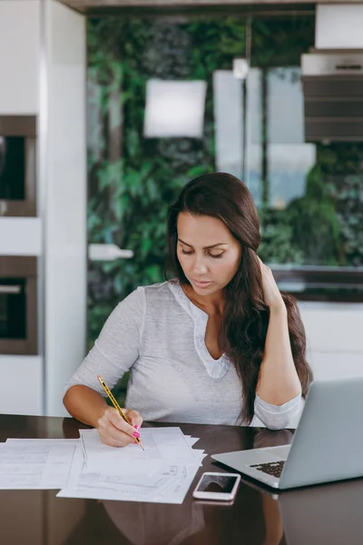 Attractive young modern business woman working with documents and laptop in the kitchen at home — Stock Photo, Image