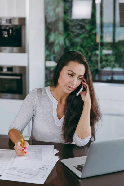Attractive young modern business woman talking on mobile phone and working with documents and laptop in the kitchen at home — Stock Photo, Image