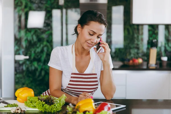 Молодая счастливая женщина в фартуке разговаривает по мобильному телефону и смотрит на рецепт в планшете на кухне. Овощной салат. Концепция диеты. Здоровый образ жизни. Готовить дома. Готовьте еду. . — стоковое фото