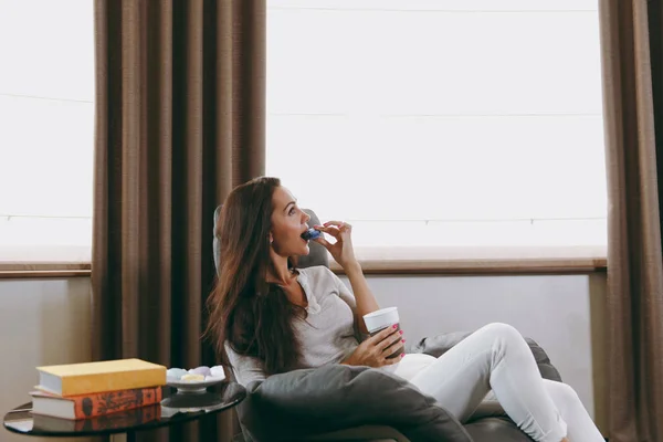 Schöne junge Frau sitzt auf modernem Stuhl vor dem Fenster zu Hause, isst Makronen und trinkt Kaffee oder Tee — Stockfoto