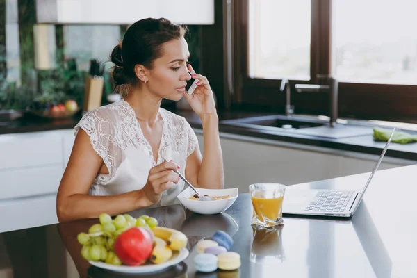 Портрет красивой счастливой женщины, разговаривающей по мобильному телефону за завтраком с ноутбуком на столе. Еда дома . — стоковое фото