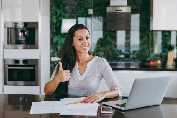 Attractive young modern business woman shows thumbs gesture cool and working with documents and laptop in the kitchen at home — Stock Photo, Image