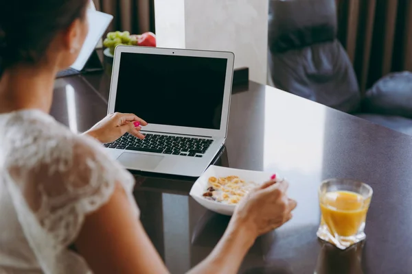 Young woman working with laptop while breakfast with cereals and milk. Eating at home close up. Back view. With place for text. — Stock Photo, Image