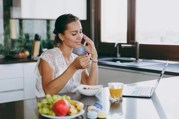 Портрет красивой счастливой женщины, разговаривающей по мобильному телефону за завтраком с ноутбуком на столе. Еда дома . — стоковое фото
