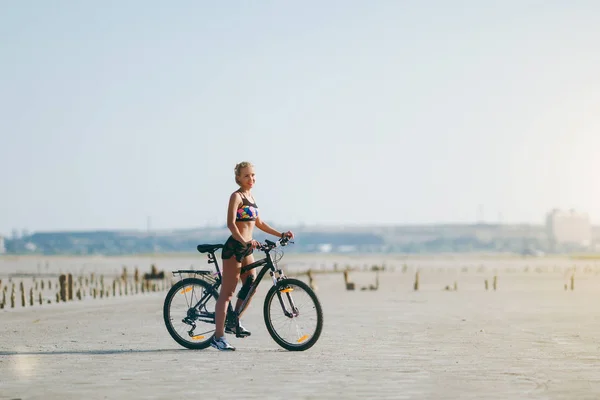 Uma mulher loira forte em um terno multicolorido se senta em uma bicicleta em uma área deserta e olha para o sol. Conceito de aptidão. Céu azul fundo — Fotografia de Stock
