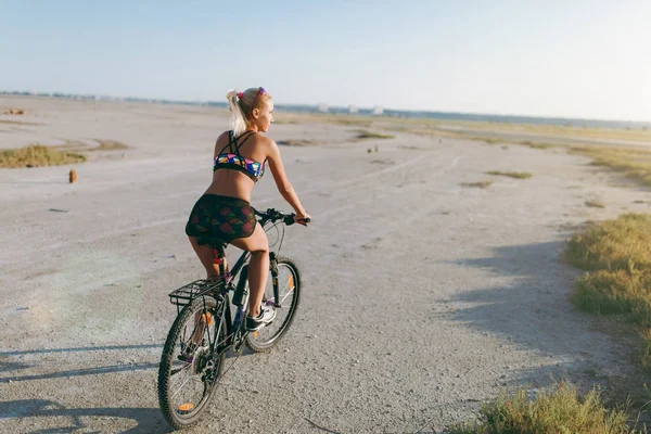 Uma mulher loira esportiva em um terno colorido monta uma bicicleta em uma deser — Fotografia de Stock