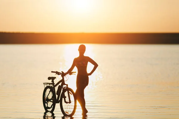 Silna Kobieta blonde w kolorowy komplet stoi w pobliżu rowerów w wodzie o zachodzie słońca na ciepły, letni dzień. Koncepcja fitness. Tło nieba — Zdjęcie stockowe