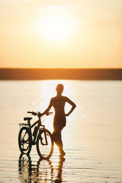 Silna Kobieta blonde w kolorowy komplet stoi w pobliżu rowerów w wodzie o zachodzie słońca na ciepły, letni dzień. Koncepcja fitness. Tło nieba — Zdjęcie stockowe