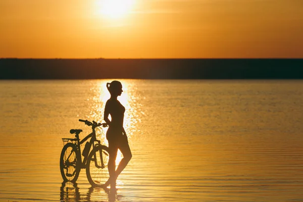 Sylwetka dziewczynki sportowy w garniturze stojącego w pobliżu rower w wodzie o zachodzie słońca na ciepły, letni dzień. Koncepcja fitness. Tło nieba — Zdjęcie stockowe
