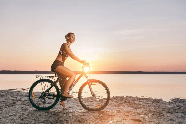 Sportowy blond kobieta w kolorowy komplet przejażdżki rowerowe w okolicy pustyni w pobliżu wody w słoneczny letni dzień. Koncepcja fitness. Na tle niebieskiego nieba — Zdjęcie stockowe