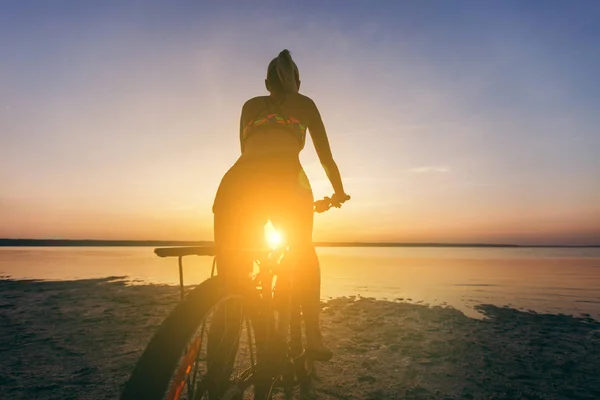 Сильная блондинка в разноцветном костюме сидит на велосипеде в пустынной местности возле воды и смотрит на солнце. Фитнес-концепция. Вид сзади — стоковое фото