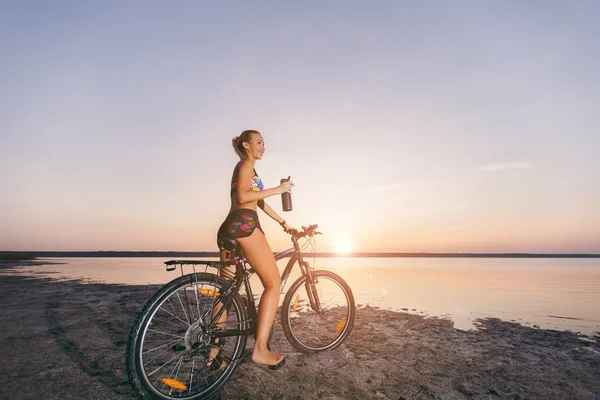 Silna Kobieta blonde w kolorowy komplet siedzi na rowerze, posiada czarne butelkę z wodą w okolicy pustyni w pobliżu wody. Koncepcja fitness. Na tle niebieskiego nieba — Zdjęcie stockowe